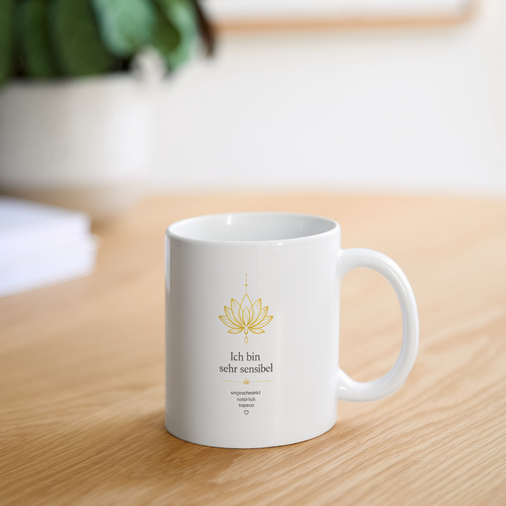Weiße Keramiktasse mit goldenem Lotusmotiv und der Aufschrift „Ich bin sehr sensibel“ auf einem Holztisch. Aura-Tasse von ungeschminkt natürlich sapeco.