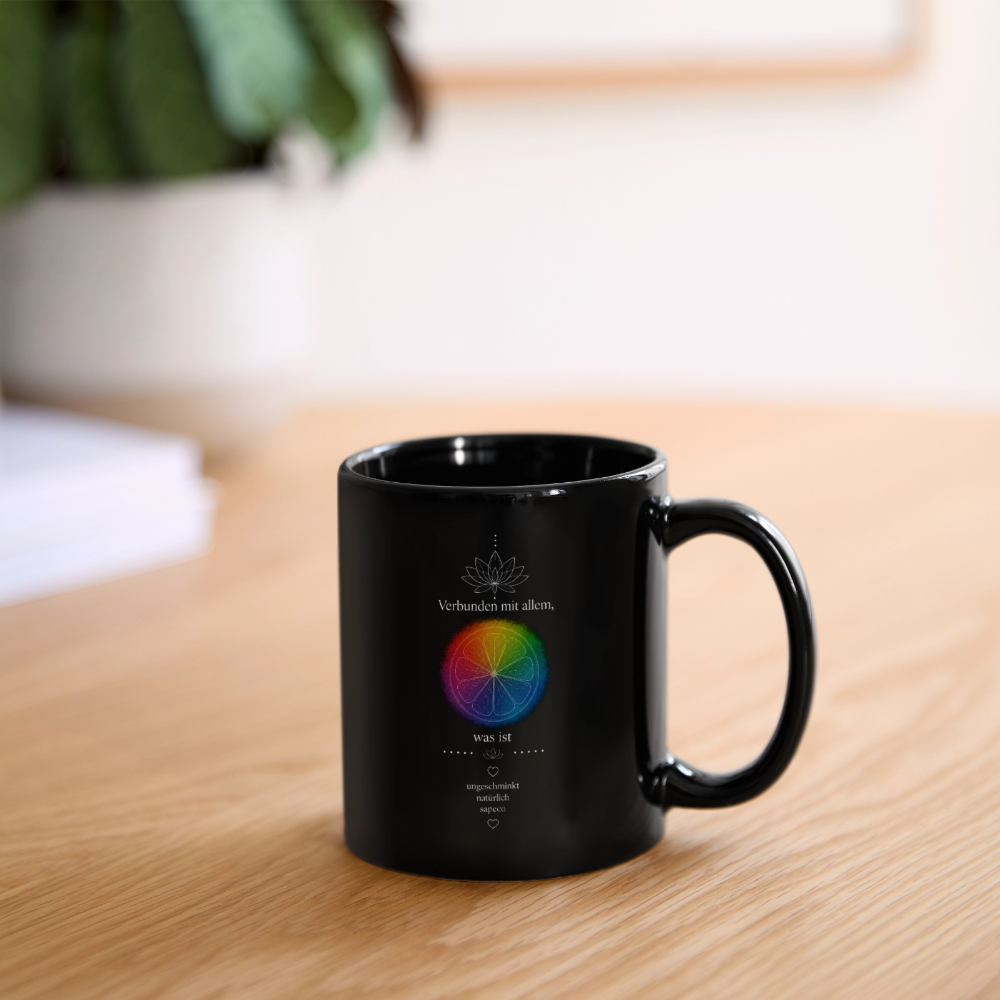 Schwarze sapeco Keramiktasse mit buntem Chakra-Motiv, Lotusblume und dem Schriftzug „Verbunden mit allem, was ist“ auf einem Holztisch in gemütlicher Wohnatmosphäre.