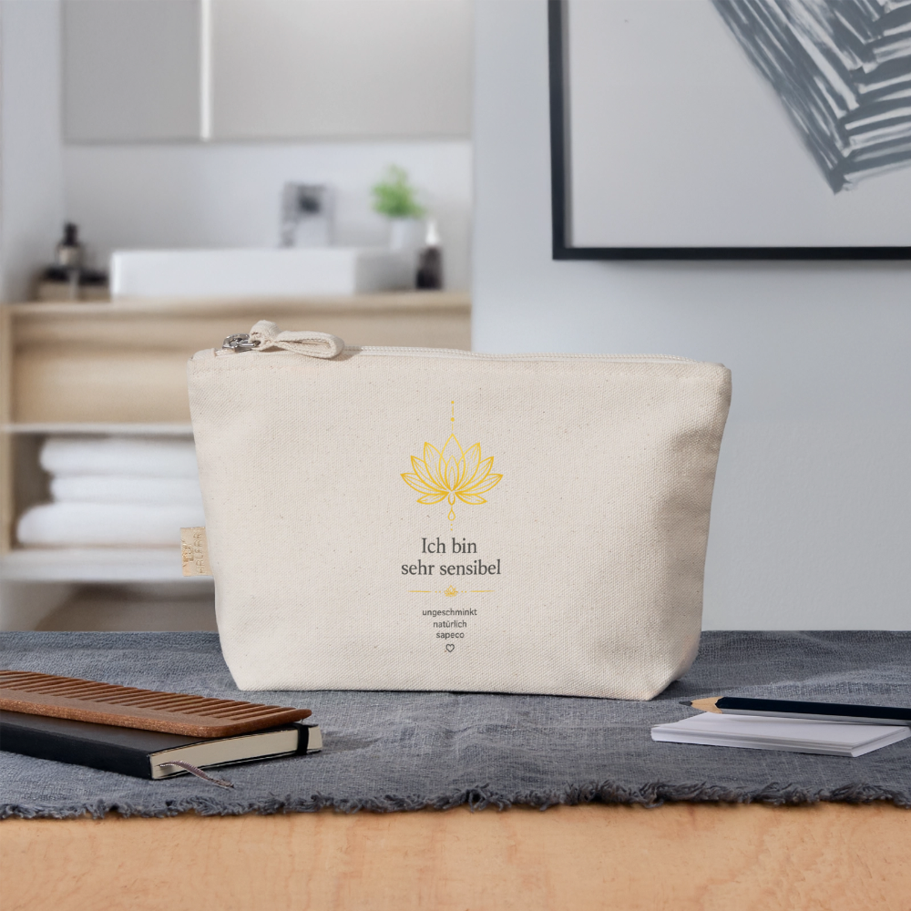 Naturfarbene sapeco Kosmetiktasche mit goldenem Lotusmotiv und Aufschrift „Ich bin sehr sensibel“ auf einem Tisch vor einem Badezimmer.