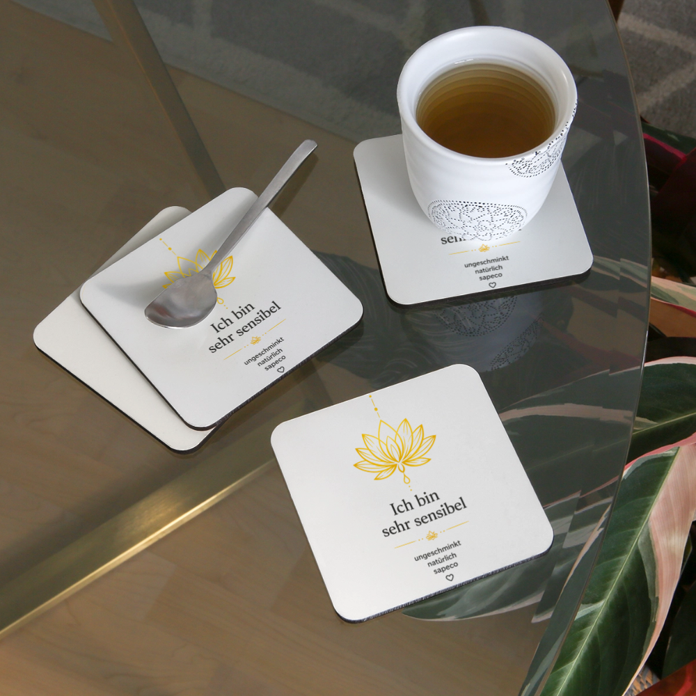 sapeco Untersetzer „Ich bin sehr sensibel“ mit goldenem Lotusmotiv und Aura-Design auf einem Glastisch. Weißes 4er-Set mit Korkrückseite, dekoriert mit Teetasse und Löffel.